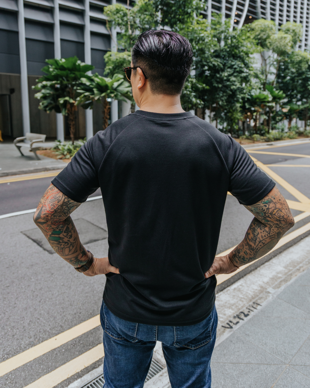 Men’s clothing for short men- Man wearing a black t-shirt and blue jeans standing on a street with trees and buildings in the background.