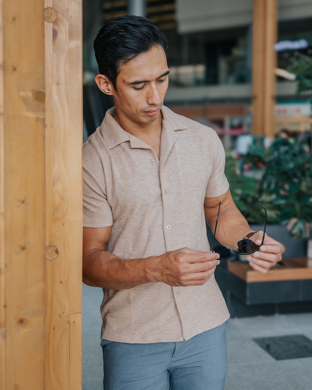 Man in a brown shirt and grey pants standing outdoors, holding sunglasses wearing clothes for men 175cm & under.