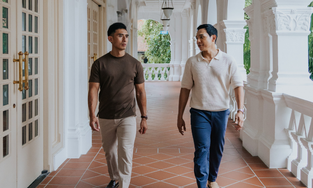 Two men walking on a tiled porch with white columns and greenery in the background. Wearing Tops for short men
