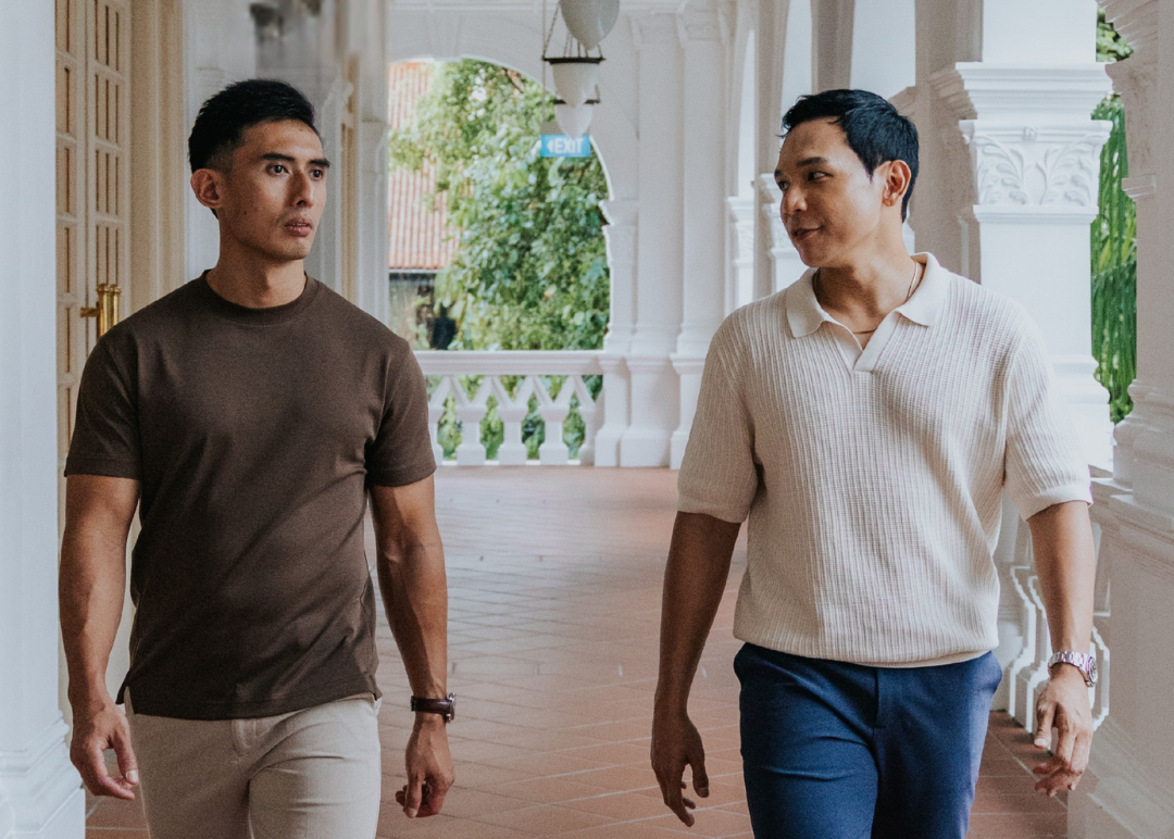 Two men walking side by side in a decorative hallway wearing a shirt and t-shirt for short men